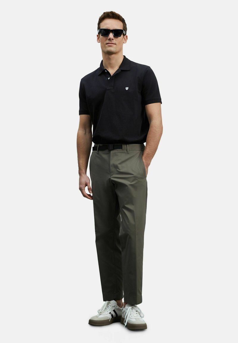 Zwart poloshirt, olijfgroene broek, zwarte riem en witte sneakers met grijze accenten. Model draagt zwarte zonnebril en staat tegen een effen achtergrond.