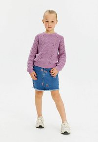 Ljust lila stickad tröja med en vågmönster, matchad med en blå denimkjol med regnbågsbroderi och fransad nederkant. Vita sneakers.