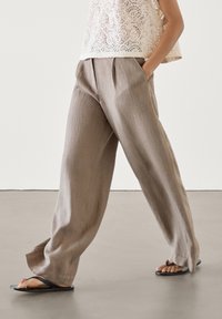 Pantalon en lin marron clair avec un devant plissé, doté de poches latérales et d'une coupe décontractée, associé à des tongs noires sur un sol en béton.