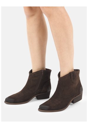 Benen met donkerbruine suède enkellaarsjes met zijritsen, lage gestapelde hakken en subtiele decoratieve stiksels op een witte achtergrond.