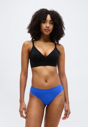 Femme portant un bralette noir et une culotte bleu taille haute, debout contre un fond clair uni, face à la caméra avec une posture détendue.