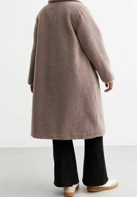 Miękki, taupe'owy płaszcz z szerokim kołnierzem i długimi rękawami, sięgający do połowy łydki. Noszony na czarnych spodniach i jasnych butach.