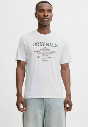 Mann trägt ein weißes T-Shirt mit dem Aufdruck "ORIGINALS 1990 Limited Edition Authentic Collection Copenhagen", kombiniert mit hellblauen Denim-Jeans.