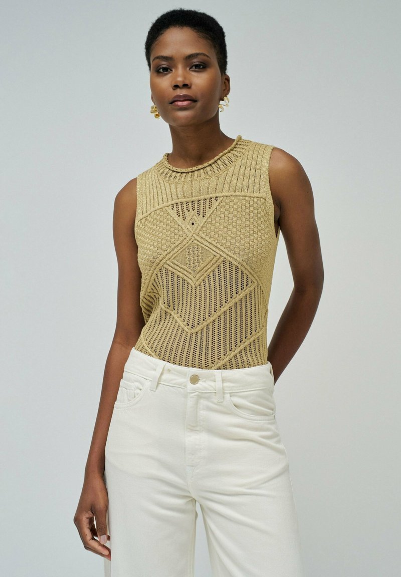 Débardeur en tricot sans manches en or clair avec des motifs en diamant complexes, associé à un jean blanc taille haute. Texture lisse et design ajusté.