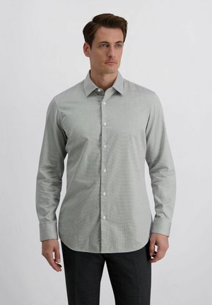 Mann trägt hellgraues langärmeliges Hemd mit Knöpfen und dunkle Hose, steht vor hellem einfarbigem Hintergrund und schaut leicht nach rechts.