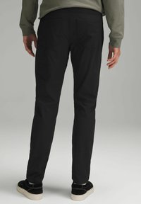 Mustad slim-fit püksid kitsenevate jalgadega, millel on kaks taga taskut ja sile tekstuur. Kandetavad mustade spordijalatsitega ja rohelise pika varrukaga särgiga.