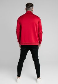 Sudadera roja con cuello alto, mangas largas y dobladillo texturizado. Combinada con vaqueros ajustados negros y zapatillas blancas.