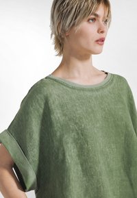 Grünes oversized Leinen-Top mit weitem Ausschnitt, hochgekrempelten kurzen Ärmeln und strukturierter Oberfläche. Dezente Farbnuancen für eine zusätzliche Tiefe.