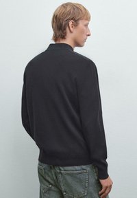 Svart stickad tröja med krage, långa ärmar och en åtsittande nederdel. Synliga sömnadsdetaljer; bärs med ljusblå denimjeans.