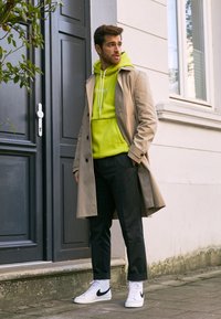 Manteau trench beige sur un hoodie vert vif avec des cordons blancs, associé à un pantalon noir et des baskets Nike blanches sur une surface pavée en extérieur.