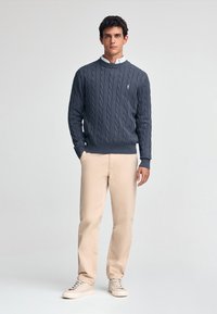 Marinblå kabelstickad tröja med rund hals, parat med ljusa beige byxor och beige sneakers. Enkelt design, mjuk textur och diskret logotyp.