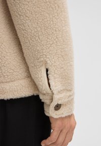 Beige fleece jacket with a textured surface, featuring a buttoned cuff and a side pocket. Soft, cozy material with a casual design.