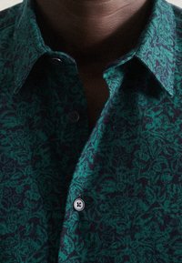 Camicia con motivo verde e navy, design floreale, texture morbida e colletto abbottonato. Presenta un unico bottone vicino al colletto.