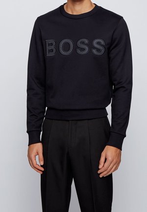 Mężczyzna w czarnej bluzie typu crewneck z wypukłym napisem "BOSS" oraz dopasowanych czarnych, szytych na miarę spodniach na jednolitym tle.
