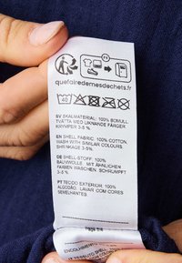 Tekstilhuvud med tvättråd på mjukt bomullstyg, vit etikett med svart text som beskriver tvättråd på flera språk, hålls framför en mörkblå bakgrund.