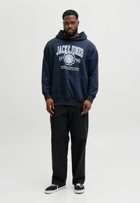 Sudadera con capucha azul marino con gran texto de logo en blanco "JACK & JONES 1990" en la parte frontal, combinada con pantalones negros y zapatillas negras.
