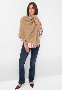 Beige knitted poncho with cable pattern draped over a white and red striped shirt. Model wears dark blue jeans and burgundy pointed slip-on shoes.