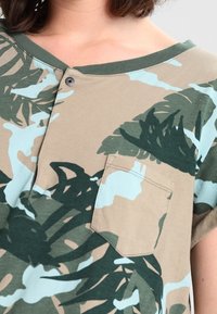 Musta lyhythihainen, taupenvärinen paita, jossa on vihreitä ja vaaleansinisiä lehtimalleja, V-kaulus, napit yksityiskohtana ja rintatasku lisäämään toiminnallisuutta.