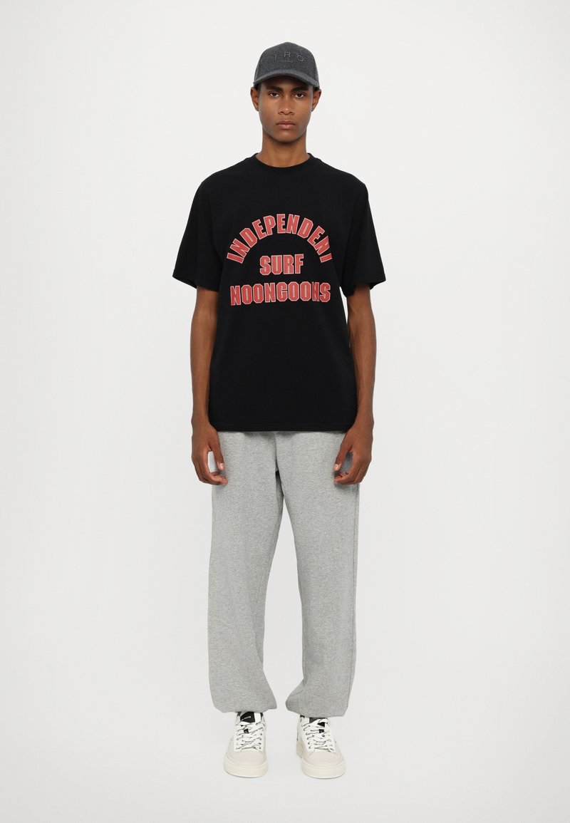 Black cotton graphic t-shirt with red text, paired with gray sweatpants. White sneakers and a dark cap complete the casual outfit.