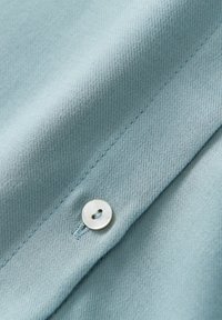 Lichtblauwe stof met een gestructureerde weving, met een ronde, glanzende knop die aan de voorkant is aangenaaid. Subtiele stikdetails zijn zichtbaar.