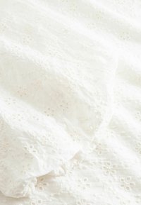 Λευκό ύφασμα με κεντητό λουλουδένιο σχέδιο με οπές και ραμμένη ελαστική άκρη, που δείχνει υφή και ελαφριές σκιές.