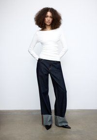 Hvid langærmet bluse med bådudskæring kombineret med mørke denimjeans med unikke detaljeringer ved kanten og frynset kanter, båret med sorte hæle.