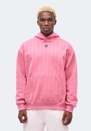 Giovane uomo con capelli biondi che indossa una felpa con cappuccio rosa con un piccolo logo sul petto e pantaloni bianchi, in piedi davanti a uno sfondo neutro.