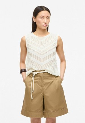 Donna che indossa un top bianco senza maniche in maglia con motivo a spina di pesce e pantaloncini marroni con cintura fino al ginocchio, mani nelle tasche, sfondo neutro.