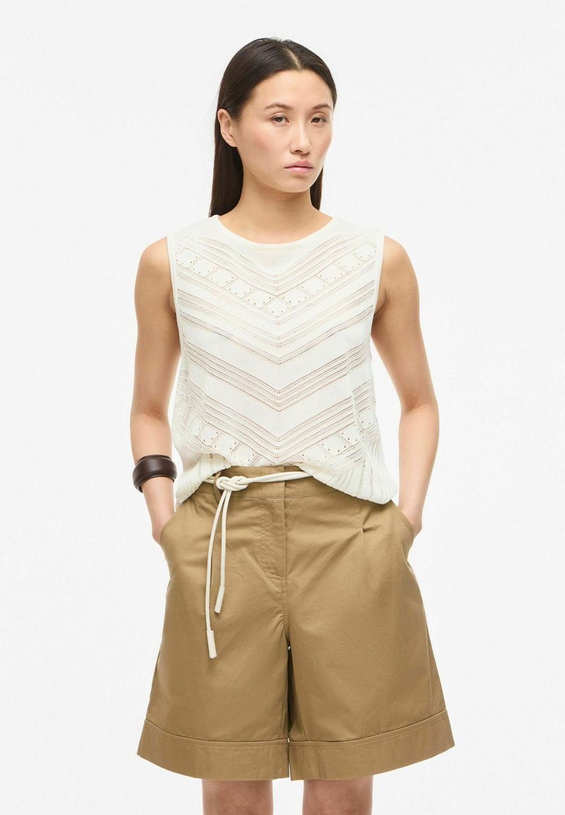 Femme portant un top en maille blanc sans manches avec motif chevron et un short beige ceinturé à hauteur des genoux, mains dans les poches, fond neutre.