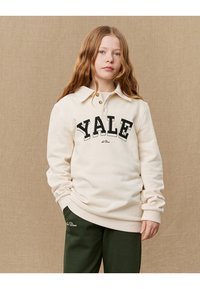Cream sweatshirt with "YALE" in bold black font, collared design, and ribbed cuffs, paired with dark green sweatpants. Textured background.