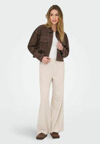 Kvinna med långt hår som bär en brun kort jacka, vit topp och beige utsvängda byxor, står framför en neutral bakgrund och ser något bortom kameran.