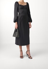 Abito in satin nero con maniche a sbuffo e scollatura quadrata, vita cintrata, lunghezza midi. Porta una piccola borsa a rete e indossa sandali neri con cinturini.