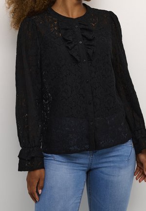 Vrouw die een zwarte kanten blouse met lange mouwen, ruchtdetail en knopen draagt, gecombineerd met lichtblauwe spijkerbroek.