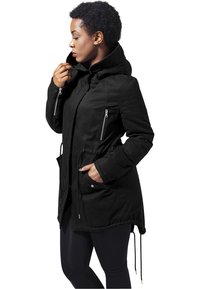 Parka noire avec capuche, poches zippées, taille ajustable par cordon et accents texturés aux épaules. Le matériau semble doux et durable.