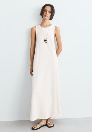 Vrouw draagt een mouwloze witte maxi-jurk, sandalen met slangenprint en een halsketting met rechthoekige hanger, staand tegen een effen lichtgrijze muur.