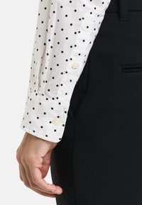 White shirt with black polka dots and a buttoned cuff, paired with black trousers. Features smooth fabric and tailored fit.
