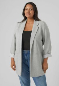 Blazer cintré gris clair à manches trois-quarts, avec des détails de poignet côtelés et deux poches avant, associé à un haut noir et un jean bleu.