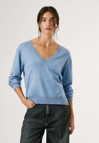 Šviesiai mėlynas V-kaklo megztinis, pagamintas iš minkštos medžiagos, su raukiniais ant rankovių ir apatinės dalies. Dėvimas su tamsiai pilkais džinsais, turi laisvą, kasdienį siluetą.