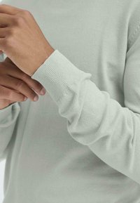 Pulover tricotat într-o nuanță verde deschis, cu o textură moale, manșete cu o bandă elastică și o croială lejeră, fără logo-uri sau modele vizibile.