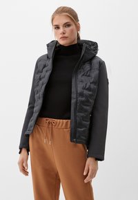 Schwarze isolierte Jacke mit gestepptem Design, angenommener Kapuze, Reißverschluss vorne und kontrastierenden glatten Ärmeln. Trägt man über einem gerippten schwarzen Oberteil und braunen Hosen.