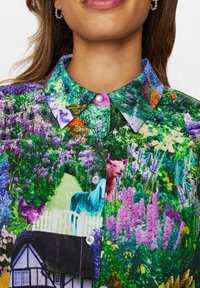 Flerfärgad skjorta med blommönster, knappad krage och bilder av djur och en stuga, tillverkad av ett mjukt tyg.