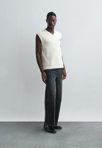 Gebreide, crèmekleurige mouwloze trui met een V-hals en een textuur van ribbels, gecombineerd met donkergrijze, recht gesneden jeans met zijnaadaccenten.