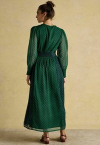 Zielona długa sukienka z marszczonym pasem, przezroczystymi długimi rękawami i diamentowym wzorem w ciemniejszej zieleni, zestawiona z brązowymi sandałami na paski.