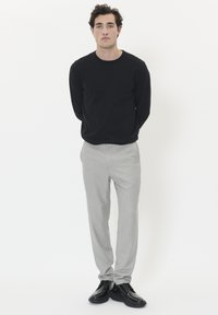 Young man standing with hands behind back wearing a black long-sleeve shirt, light grey pants, and black dress shoes on white background.
