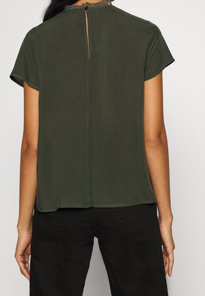 Kobieta ubrana w ciemnozieloną bluzkę z krótkim rękawem, wykończoną koronką i zapinaną na guziki z tylu z wycięciem w kształcie klucza, w połączeniu z czarnymi spodniami.