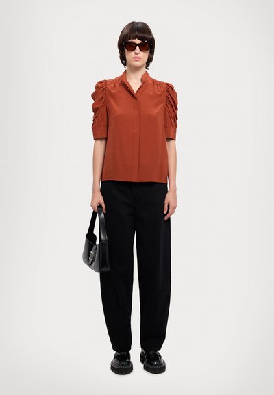Blusa cor ferrugem com mangas bufantes, combinada com calças largas pretas e sapatos pretos. Uma bolsa preta e óculos de sol completam o visual.