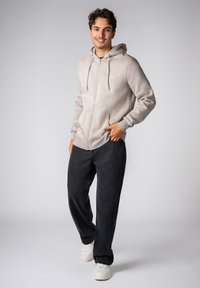 Hellgrauer Kapuzenpullover mit Reißverschluss, Kordeln, Fronttaschen und Logodetail. Kombiniert mit locker sitzenden schwarzen Jeans und weißen Turnschuhen.