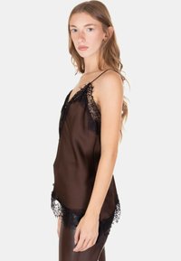 Giovane donna con lunghi capelli chiari che indossa una camisola nera trasparente bordata di pizzo e pantaloni scuri, in piedi di lato contro uno sfondo neutro.