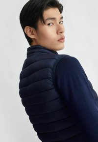 Gilet imbottito navy con cuciture orizzontali, colletto alto e design senza maniche, abbinato a una camicia navy a maniche lunghe sotto.