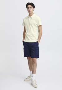 Lys gul polo-skjorte med teksturert overflate, kombinert med marineblå cargo-shorts. Modellen har på seg hvite joggesko og hvite sokker. Enkelt bakgrunn.